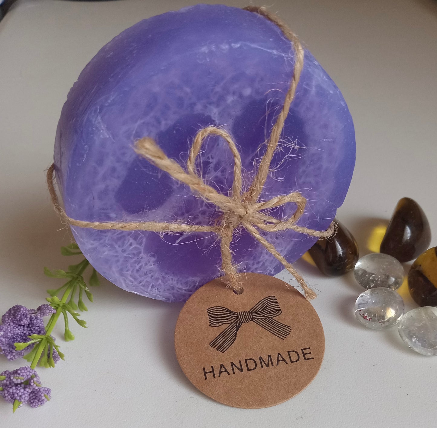 Zaya Soaps Loofah Soaps -Round handmade purple soap tied with twine bow and thank you tag displayed with decorative stones By Zaya Soaps LLC